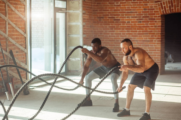 Twee multi-etnisch blote borst sporters trainen met de slag bij touwen op de sportschool. - Foto, afbeelding