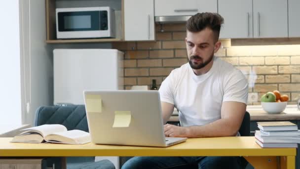 El hombre aprende y escribe el trabajo en una computadora en proyecto usando un libro de texto sentado en la cocina en casa
. - Metraje, vídeo