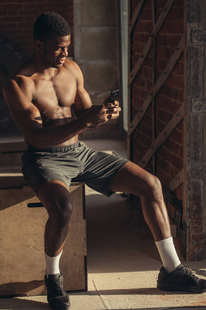 Shirtless african american athlete resting and using smartphone at gym - 写真・画像