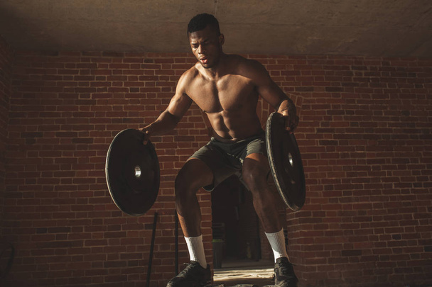 Shirtless african man with barbell plates jumps in air on truck tire in gym - Foto, Imagem
