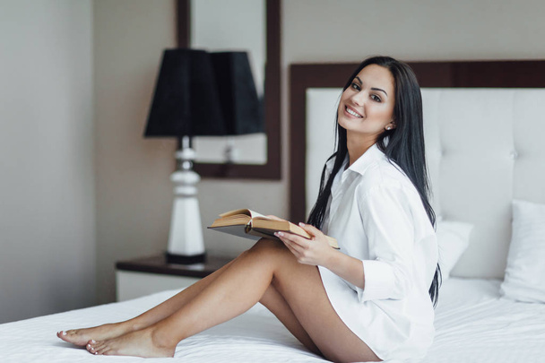 Portrait of happy brunette woman sitting in bed reading book  - Fotó, kép