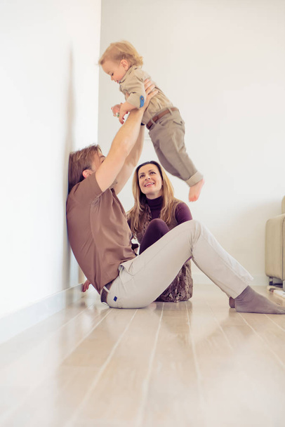 Happy family sitting on floor with their little baby. Family spending time at home with their son. Father raise his child in the air. - Φωτογραφία, εικόνα