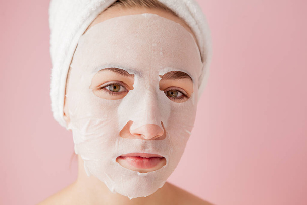 Beautiful young woman is applying a cosmetic tissue mask on a face on a pink background. Healthcare and beauty treatment and technology concept - Foto, imagen