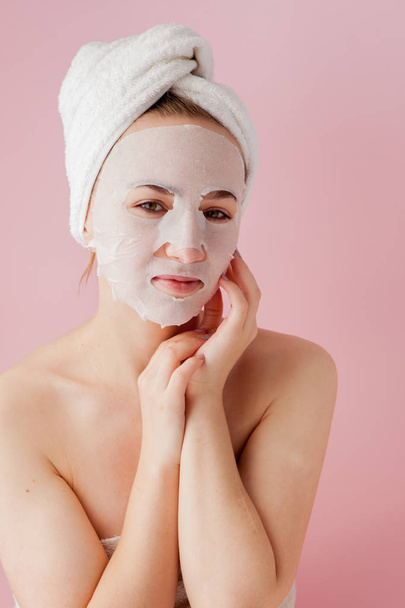 Beautiful young woman is applying a cosmetic tissue mask on a face on a pink background - Foto, afbeelding