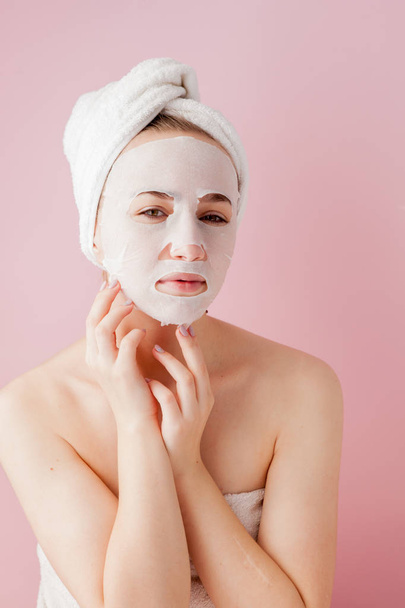 Beautiful young woman is applying a cosmetic tissue mask on a face on a pink background. Healthcare and beauty treatment and technology concept - Foto, Imagen