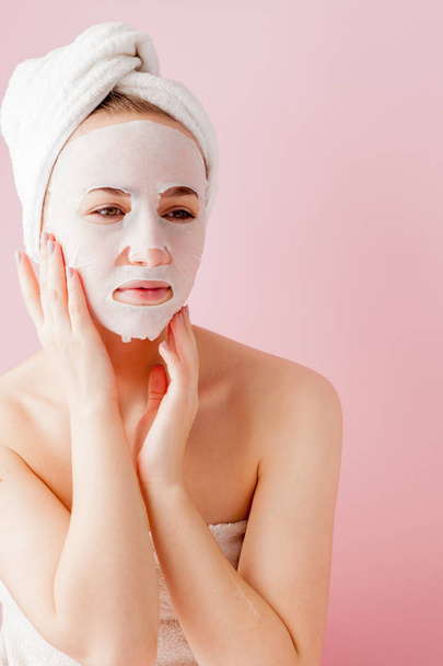 Beautiful young woman is applying a cosmetic tissue mask on a face on a pink background. Healthcare and beauty treatment and technology concept - Valokuva, kuva
