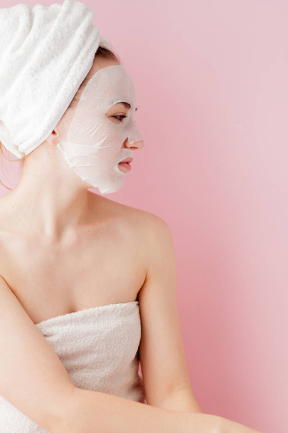 Beautiful young woman is applying a cosmetic tissue mask on a face on a pink background. Healthcare and beauty treatment and technology concept - Φωτογραφία, εικόνα