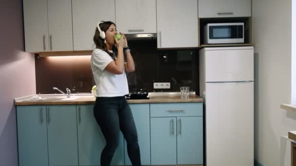 Happy woman dancing and listening to music with headphones in the kitchen cooking and eating apple. - Video