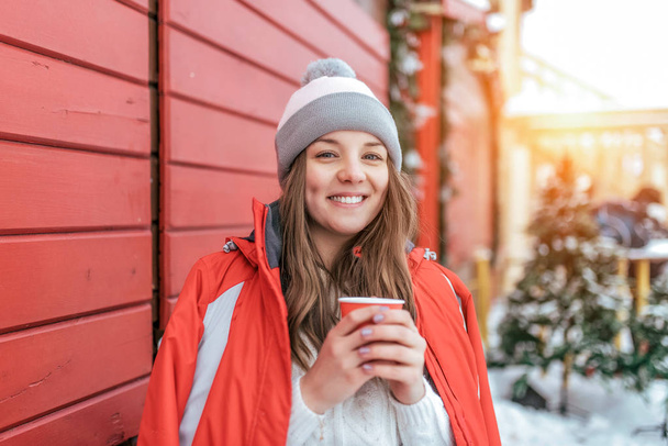 Menina bonita no inverno em uma jaqueta no fundo de estores de madeira vermelhos. A segurar uma chávena de chá de café. Feliz sorrir e rir. Resort de férias no fim de semana
. - Foto, Imagem