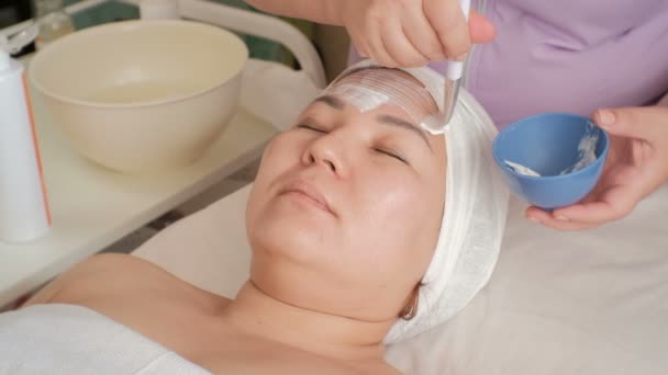 Die Kosmetikerin trägt weiße Massagecreme mit einem Pinsel auf das Gesicht einer Frau mittleren Alters auf. asiatische Mädchen in einem Schönheitssalon mit geschlossenen Augen. der Prozess der Verjüngung, des Liftens und der Entspannung - Filmmaterial, Video