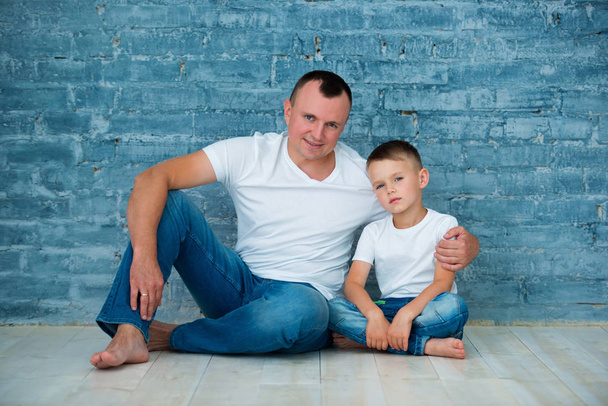 Father and son in casual jeans and white T-shirts are sitting on a warm wooden floor near a gray brick wall background - Photo, image