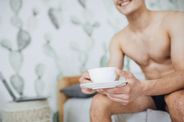 young handsome man sitting in bed in room and drinking coffee in morning - Фото, зображення