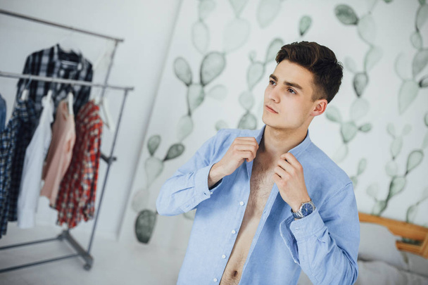Handsome young man buttoning white shirt while getting dressed at home - Foto, afbeelding