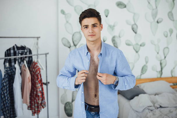 Handsome young man buttoning white shirt while getting dressed at home - Фото, зображення