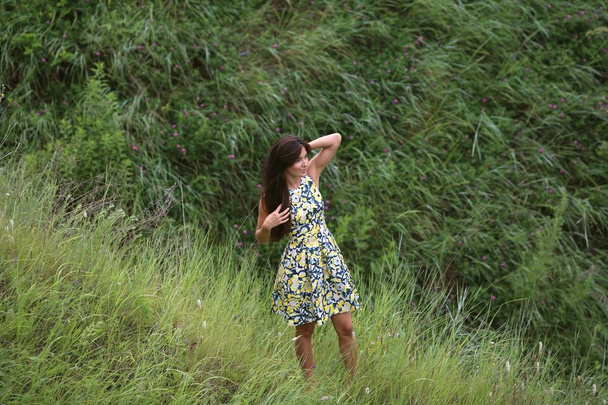 summer walk on a green ravine, a young slim pretty girl with long brown hair in a yellow dress sundress, enjoys life on the field with summer flowers - Foto, Bild