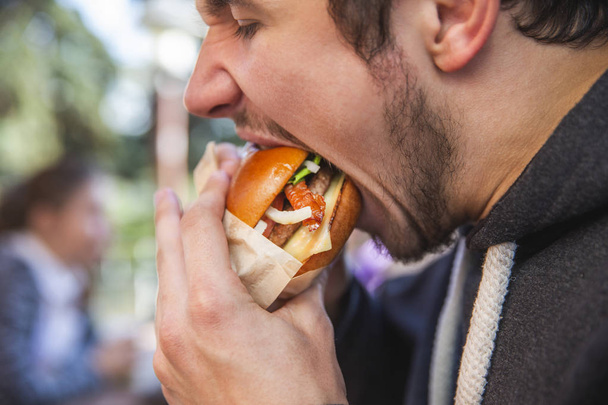 Задоволений молодий чоловік з жадібністю їсть бургер, очі закриті. Він сидить на фастфуд терасі
. - Фото, зображення