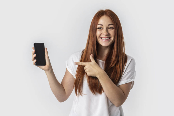 Young red-haired girl isolated on gray background. Shows a finger on the blank screen of the phone - Photo, image