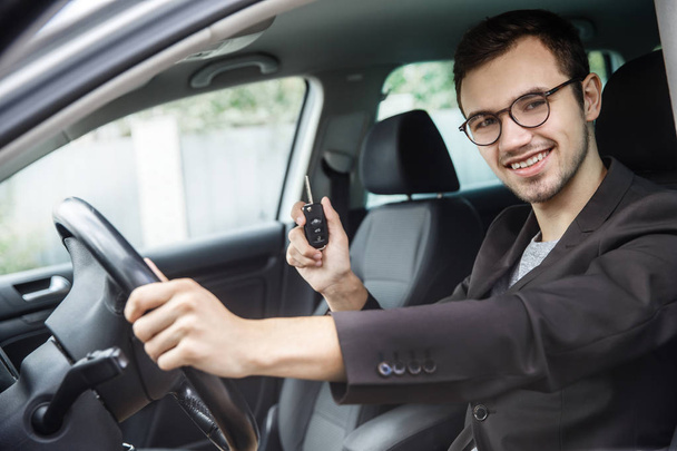 Νεαρός άνδρας κάθεται στο αυτοκίνητό του ενώ κοιτάζοντας την κάμερα. Κρατάει τα κλειδιά στο δεξί του χέρι. Το αριστερό του χέρι είναι στο τιμόνι. - Φωτογραφία, εικόνα