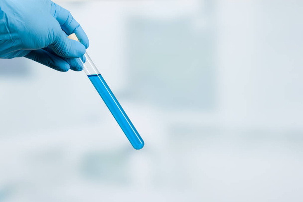 hand in gloves holds a test tube with blue liquid in front of a laboratory background - 写真・画像