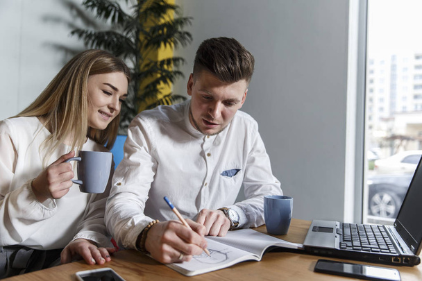 Young man and woman are sitting together at table and looking at journal. Guy is writing down. There is a cup and laptop on table. They are concentrated. Guy and girl are working. - Фото, зображення
