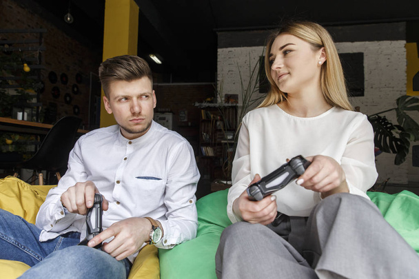 Young man and woman are sitting together on soft chairs and playing on gamepads. They look at each other and are resting. - Fotografie, Obrázek