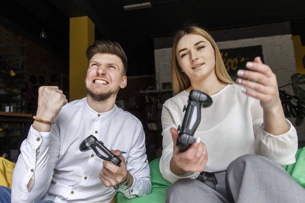 Young man and woman are sitting together on soft chairs and playing on gamepads. Guy feels the victory. Nice girl is sitting and looks straight. They are resting. - Photo, Image