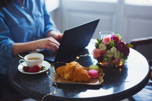 Jovem freelancer em camisa azul trabalhando no laptop durante o almoço com café, croissant, macaroons e flores na mesa à luz do dia
 - Foto, Imagem
