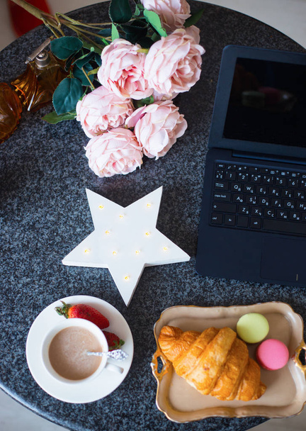 Lente plat lag met kopje latte, witte Led star, laptop, pioenrozen, croissant en bitterkoekjes op donkere marmeren tafel - Foto, afbeelding