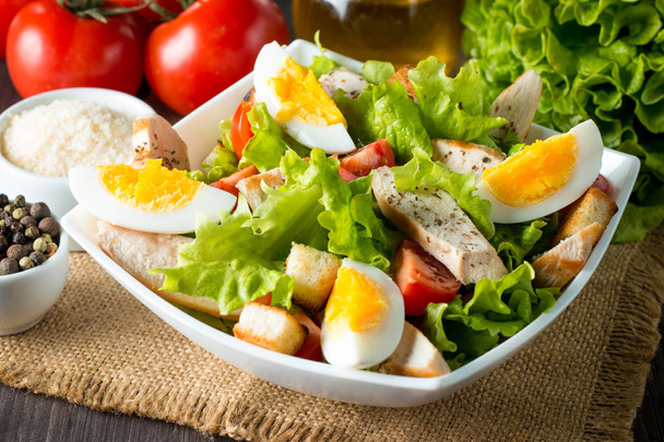 Φρέσκια σαλάτα από ντομάτα, ρόκα, στήθος κοτόπουλου, αυγά, ρόκα, κράκερ και μπαχαρικά. Σαλάτα του Καίσαρα σε ένα λευκό, διάφανο μπολ σε φόντο ξύλινη - Φωτογραφία, εικόνα