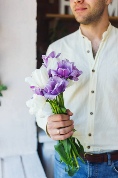 Young male hands holding spring bouquet of purple and white tulips near window in daylight. - Photo, image