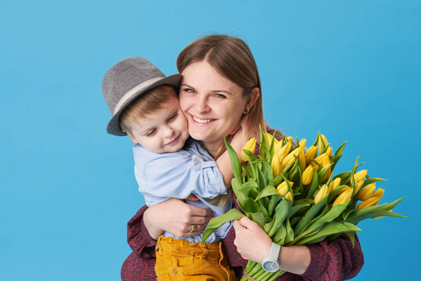 Young mother hugs his little son sitting on the floor against a blue background. Nearby lies a bouquet of spring yellow flowers. Care and relationships and family concept. Mothers day - Фото, зображення