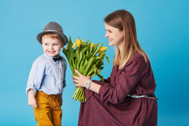 Young mother hugs his little son sitting on the floor against a blue background. Nearby lies a bouquet of spring yellow flowers. Care and relationships and family concept. Mothers day - Foto, Imagen