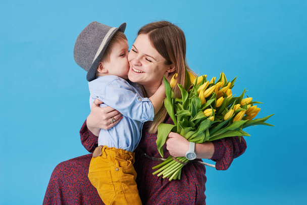 Fiatal anya hugs a kis fia, ült a földön, a kék háttér. A közelben fekszik egy csokor sárga tavaszi virágok. Ellátás és a kapcsolatok és a család fogalmát. Anyák napja - Fotó, kép