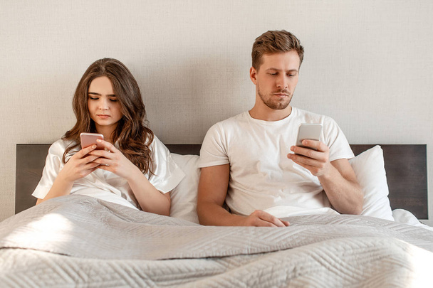 Young couple in the bed. An attractive woman and man are checking social networks news in mobile phones, texting or sending messages - 写真・画像