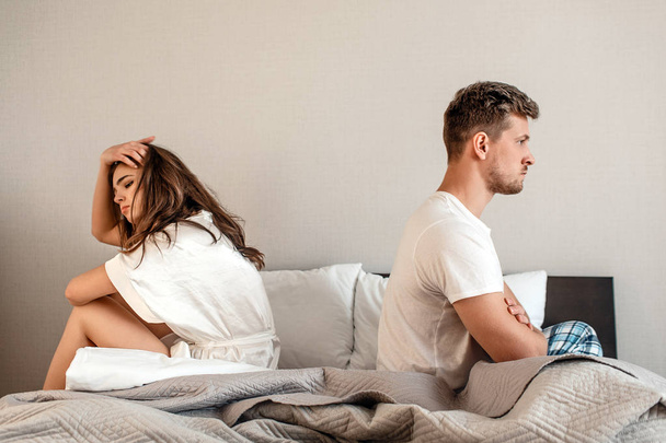 Young couple in the bedroom. Unhappy man and woman are sitting back to back in the bed, couple quarrel home scene - Fotoğraf, Görsel