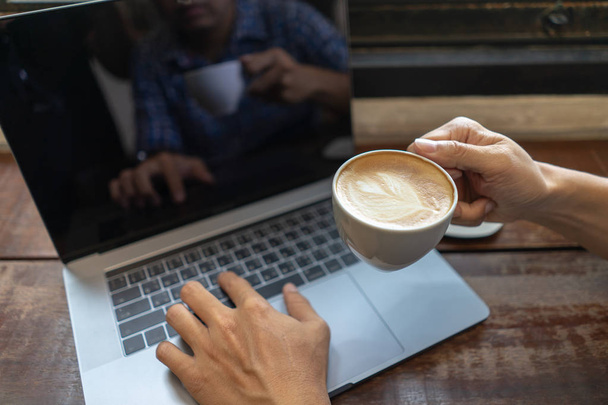 Business man holding coffee cup and typing on laptop computer keyboard on wooden table. Close up male hands with notebook pc drinking coffee cup, business, education, people and technology concept. - Photo, image