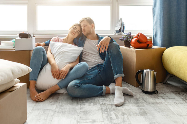 Young family couple bought or rented their first small apartment. People sit on floor and sleep. Tired and exhausted. Moving unpacking. Young woman hold white pillow. - Foto, imagen