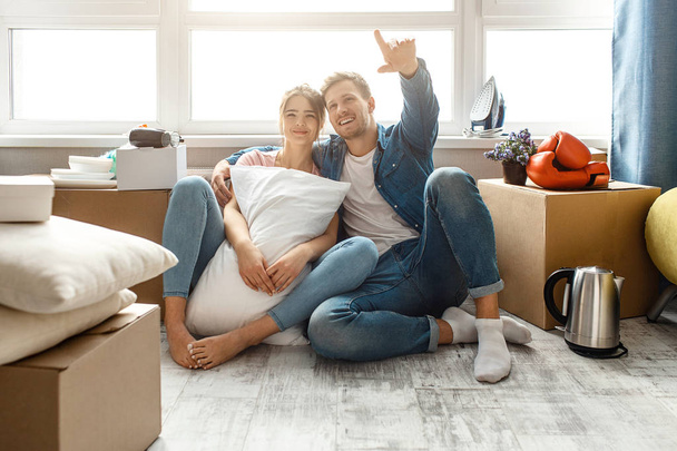 Young family couple bought or rented their first small apartment. Lovely people sit on floor and look up forward. Guy point. He hugs young woman. Moving in and unpacking. - Foto, Imagen