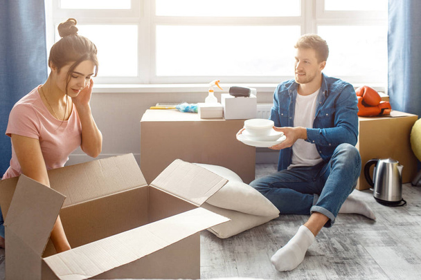 Young family couple bought or rented their first small apartment. Guy sit on floor and hold white plates. Young woman look into box. They unpack stuff in room. Moving in. - Foto, Imagen
