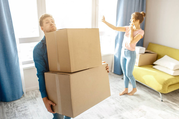 Young family couple bought or rented their first small apartment. Guy carry heavy boxes alone. Young woman stand at window and take selfie. Moving in. Unpacking. - Φωτογραφία, εικόνα
