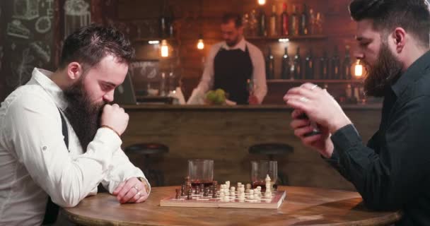 Twee bebaarde vrienden drinken en roken tijdens het spelen van schaken in een pub - Video