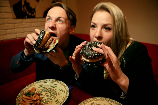 The guy and the girl eat cheeseburger with black bread, beef patty, bacon, tomatoes and pieces of cheese, dressed with sauce and green salad, funny portrait - Photo, Image