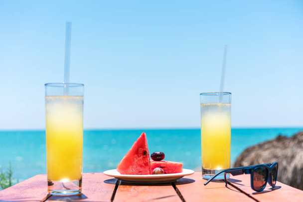 Two glasses of fresh tropical fruit juice on the beach, and a plate with grapes and watermelon on the table, against the sea. Holiday and vacation concept - Foto, Bild