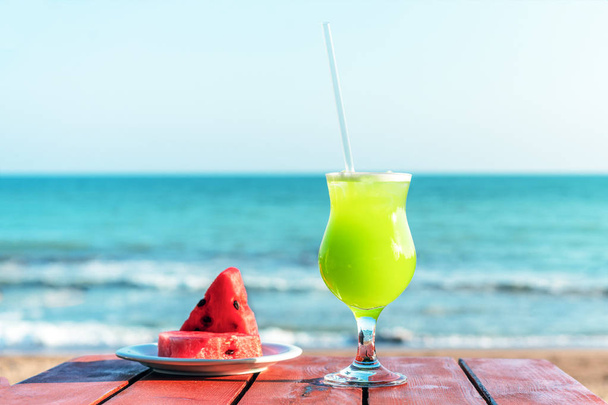 a glass of fresh green tropical fruit juice on the beach, and a plate with watermelon on the table, against the sea. The concept of summer holidays and vacation - Valokuva, kuva