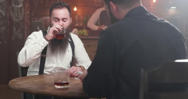 Twee gebaarde mannen in een bar drinken, socialiseren en beginnen met het spelen van een kaartspel - Video