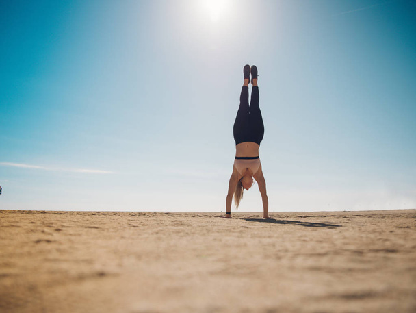 athletic woman in sportswear standing on hands on beach - Photo, Image