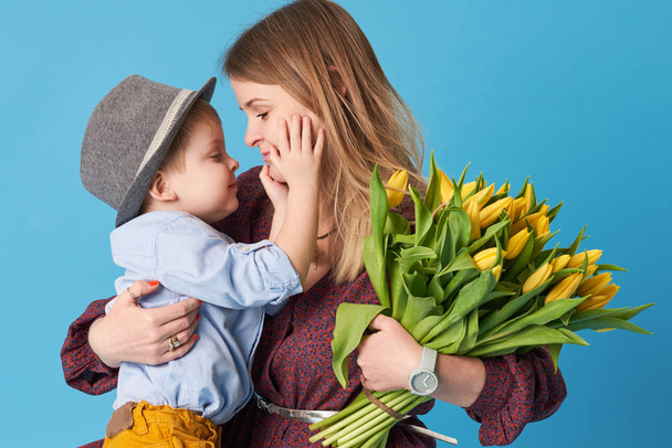 Fiatal anya hugs a kis fia, ült a földön, a kék háttér. A közelben fekszik egy csokor sárga tavaszi virágok. Ellátás és a kapcsolatok és a család fogalmát. Anyák napja - Fotó, kép