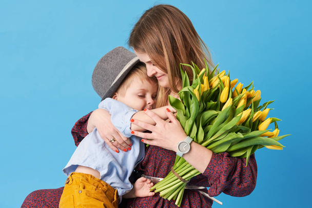 Genç anne küçük oğlu mavi bir arka plan karşı katta oturan sarılmak. Yakın bir buket bahar sarı çiçek yatıyor. Bakım ve ilişkiler ve aile kavramı. Anneler günü - Fotoğraf, Görsel