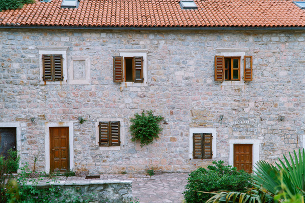 Παλιό ευρωπαϊκό οικοδόμημα με κόκκινο πλακίδιο στρογγυλό παράθυρο με ξύλινη πόρτα. Ένας θάμνος που μεγάλωσε σε ένα πέτρινο τοίχο. - Φωτογραφία, εικόνα