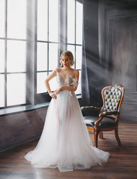 charming excellent lady became bride, girl with blond gathered hair tries on wedding chic white luxurious light dress in spacious room with large windows and bright light, naked gentle shoulder - Photo, Image